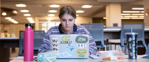 ϲʿֱֳ student studying on laptop inside the library at table with water bottle next to her